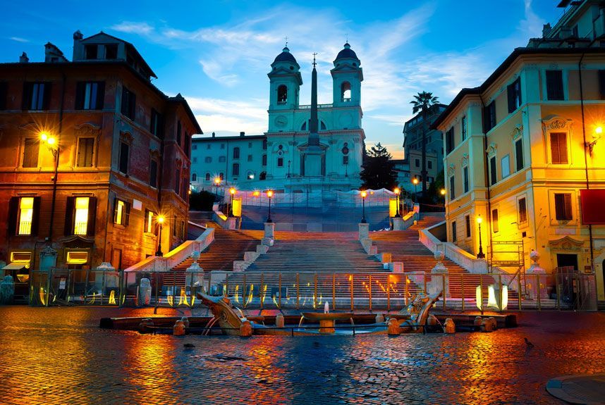 Plaza de España en Roma al anochecer, iluminada por las farolas. Los edificios enmarcan las escaleras que conducen a una iglesia con dos campanarios, reflejándose en el pavimento mojado.
