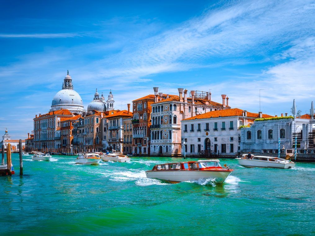 Escena del canal de Venecia con barcos pasando por edificios coloridos, una gran cúpula y un cielo azul brillante.
