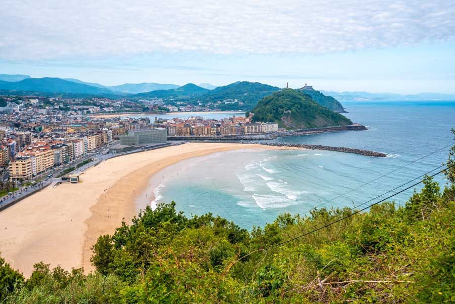 Vista panorámica de San Sebastián, España. Una playa de arena se extiende a lo largo de la costa, con la ciudad y una colina verde al fondo bajo un cielo nublado.