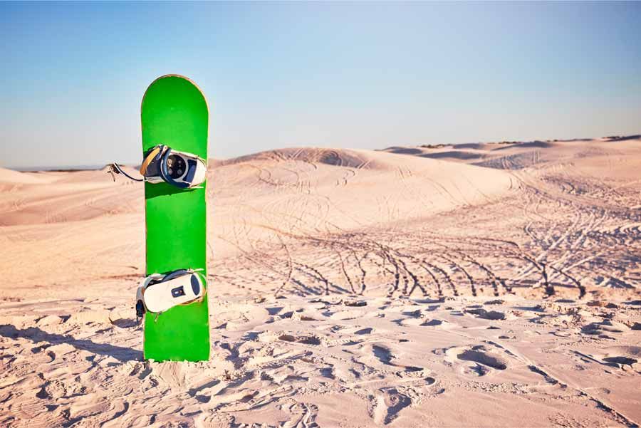 Tabla de snowboard verde brillante en un paisaje desértico arenoso. La tabla está en posición vertical, mirando a la cámara, con dunas de arena al fondo.
