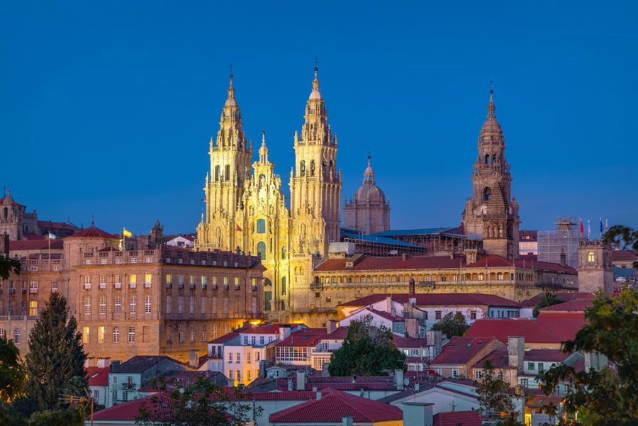 La Catedral de Santiago de Compostela iluminada al anochecer, elevándose sobre la ciudad con sus agujas ornamentadas y sus edificios de piedra de tonos cálidos.