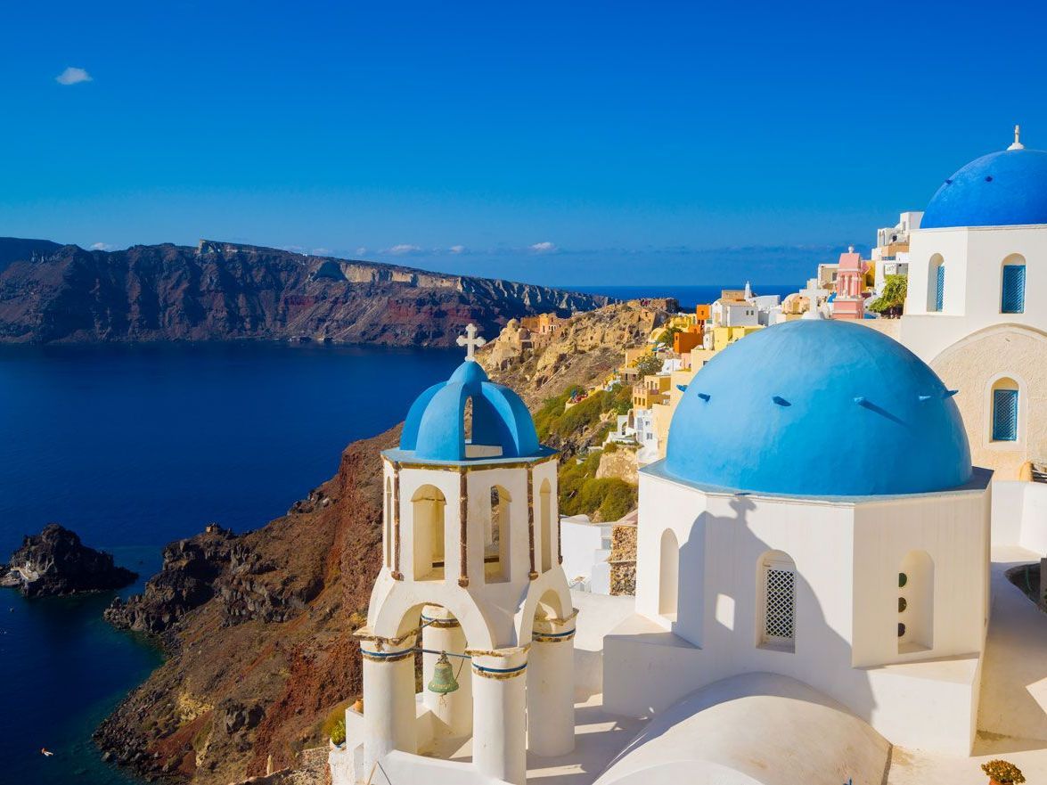 Edificios blancos con cúpulas azules y un campanario en un acantilado con vistas al mar Egeo en Santorini, Grecia.