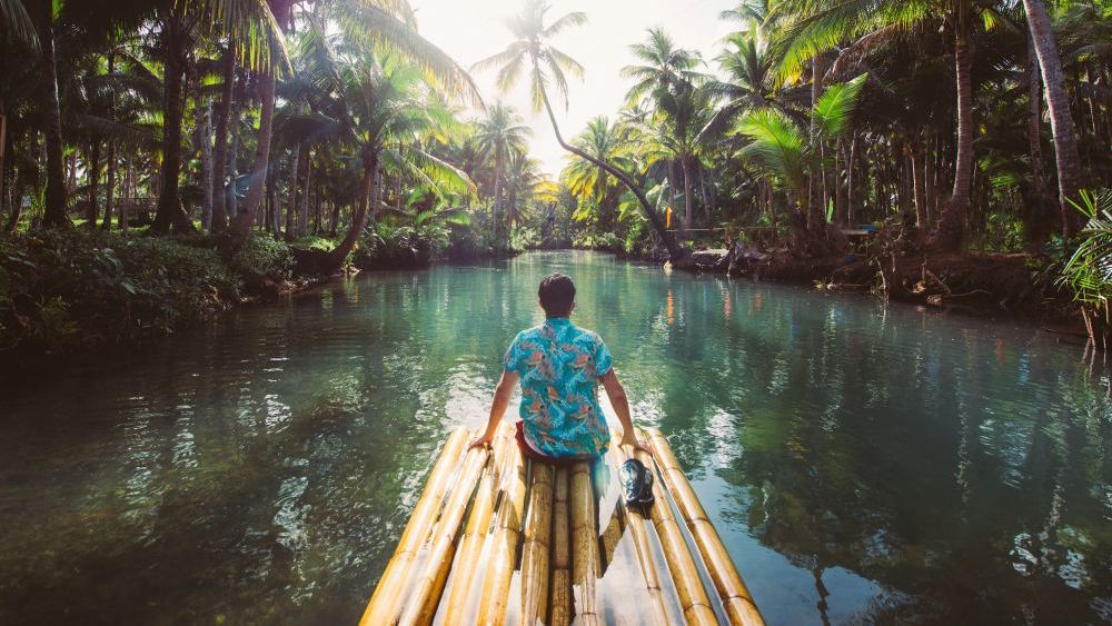 Persona en una balsa de bambú en un río turquesa, rodeada de exuberantes árboles verdes y palmeras bajo un cielo soleado.