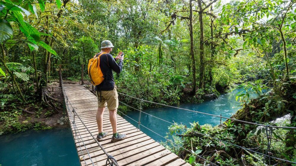 Hombre tomando una foto en un puente de madera sobre aguas turquesas en una exuberante selva tropical.