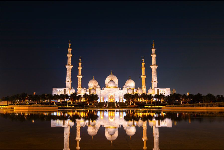 La Gran Mezquita Sheikh Zayed, iluminada por la noche, se refleja en las tranquilas aguas. Presenta cúpulas blancas, minaretes y una cálida iluminación que contrasta con el cielo oscuro.