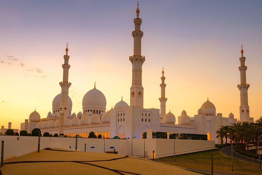 Una gran mezquita blanca con múltiples cúpulas y minaretes frente a una colorida puesta de sol en Abu Dhabi.