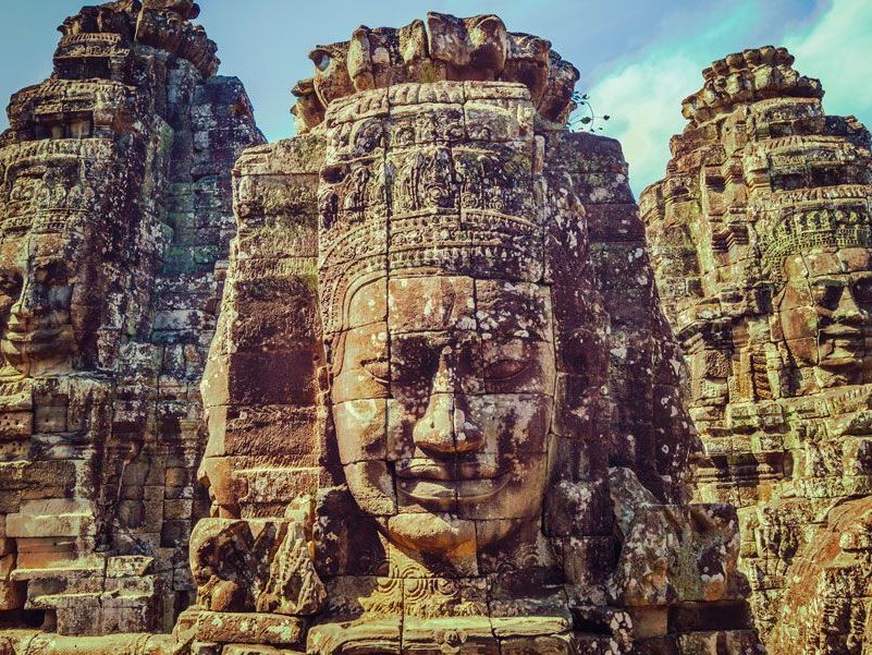 Tallas faciales en piedra en el Templo Bayón, Angkor, Camboya. Rostros con expresiones serenas emergen de las torres, de un marrón erosionado por el tiempo, contra un cielo brillante.