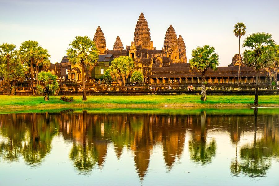 El templo de Angkor Wat reflejado en un lago tranquilo, con césped verde, árboles y un suave cielo al atardecer.