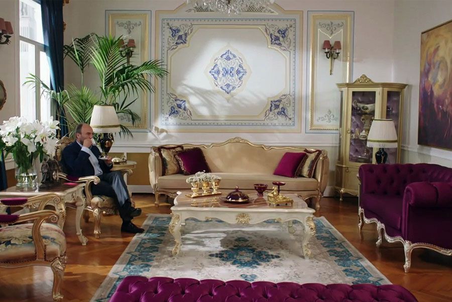 Hombre sentado en una lujosa sala de estar, con muebles ornamentados en dorado y burdeos. Se ven una alfombra decorativa y un panel de pared con diseños intrincados.