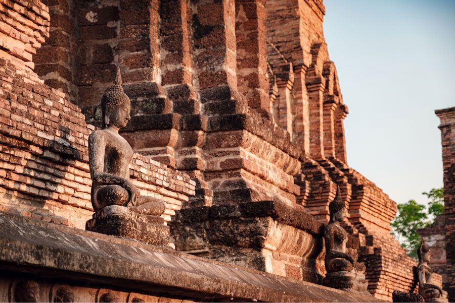 Estatuas de Buda de piedra se alinean en una pared de ladrillos desgastados en un entorno al aire libre, bañadas por la cálida luz del sol.