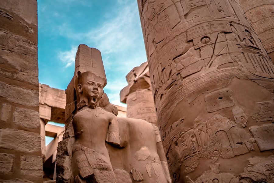 Estatua de un faraón con el rostro dañado, entre grandes columnas de piedra con jeroglíficos, bajo un cielo azul.