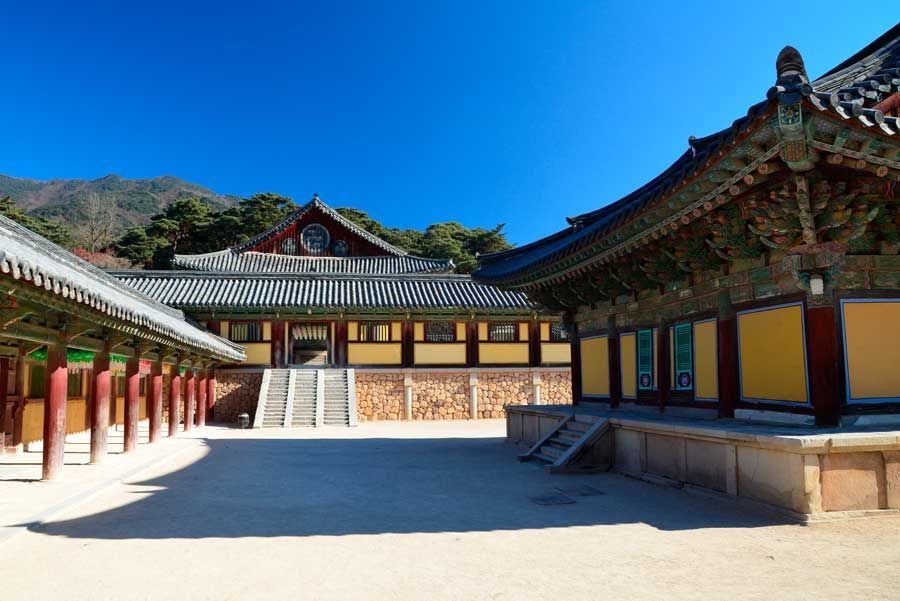 Un patio de un templo tradicional coreano con edificios coloridos y ornamentados y un fondo de montañas bajo un cielo azul claro.