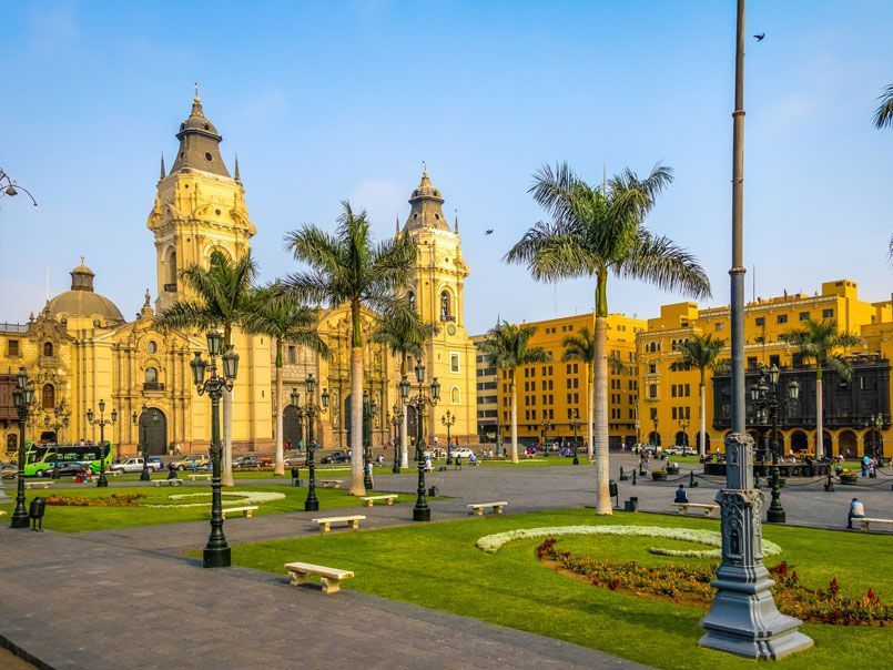 Una vista soleada de la Plaza Mayor de Lima, Perú, con una gran catedral amarilla, palmeras y césped verde.