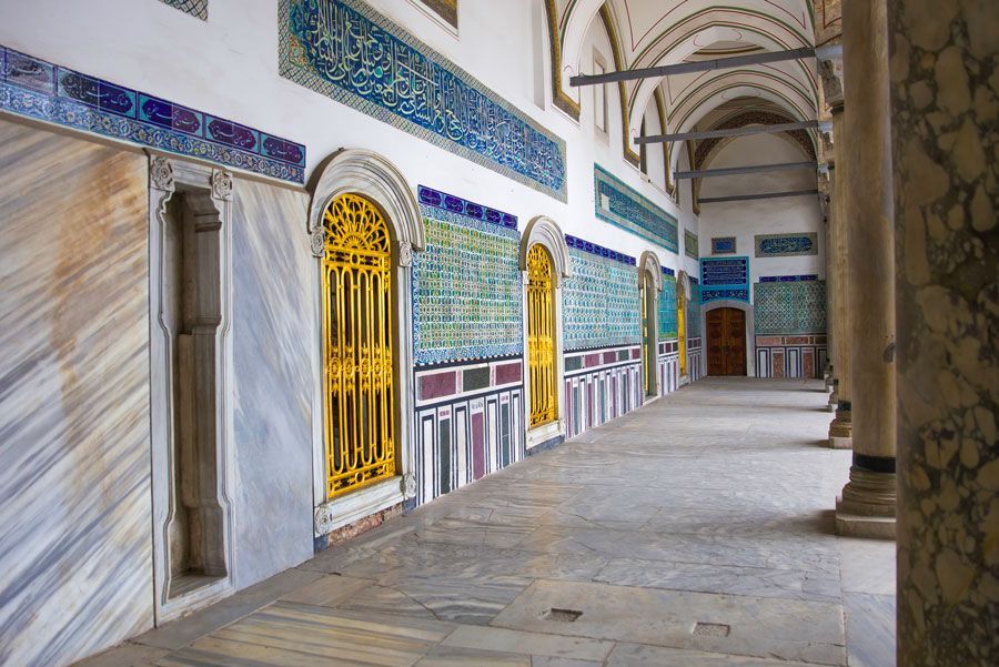 Largo pasillo en el Palacio de Topkapi, Estambul, con paredes y columnas de mármol, puertas doradas y azulejos turquesas con escritura árabe.