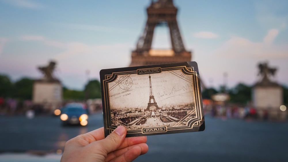 Mano sosteniendo una postal con un dibujo vintage de la Torre Eiffel, con la Torre Eiffel real y un puente en el fondo borroso.