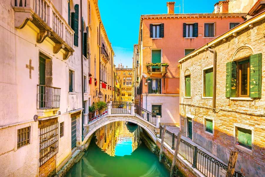 Un estrecho canal en Venecia, Italia, con coloridos edificios a lo largo del canal. Un pequeño puente arqueado lo cruza.