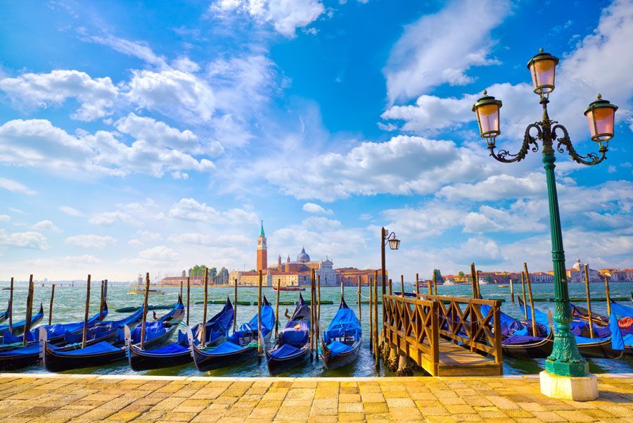 Paseo marítimo de Venecia con góndolas, un muelle de madera y una iglesia a lo lejos, bajo un cielo azul con nubes. Una farola se alza en primer plano.