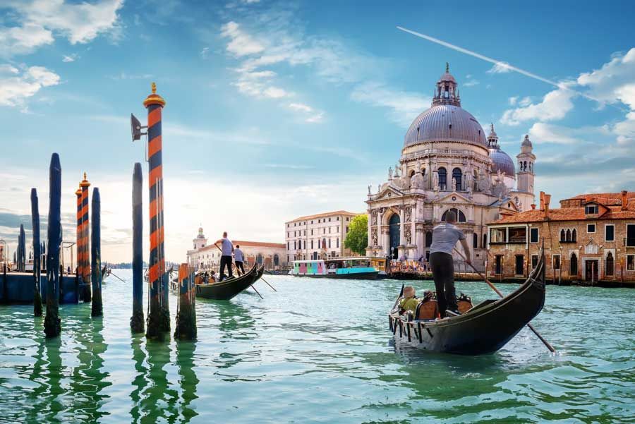 Góndolas en un canal de Venecia, con una gran iglesia con cúpula al fondo bajo un cielo azul.
