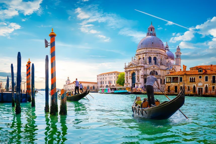 Las góndolas se deslizan sobre aguas turquesas en Venecia, Italia, con una gran iglesia con cúpula al fondo y edificios coloridos.