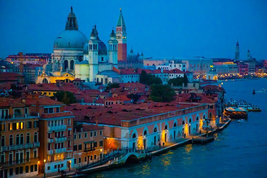 Venecia, Italia, al atardecer, con edificios a orillas del agua. Al fondo, una gran iglesia con cúpula y una torre, y luces en los edificios.