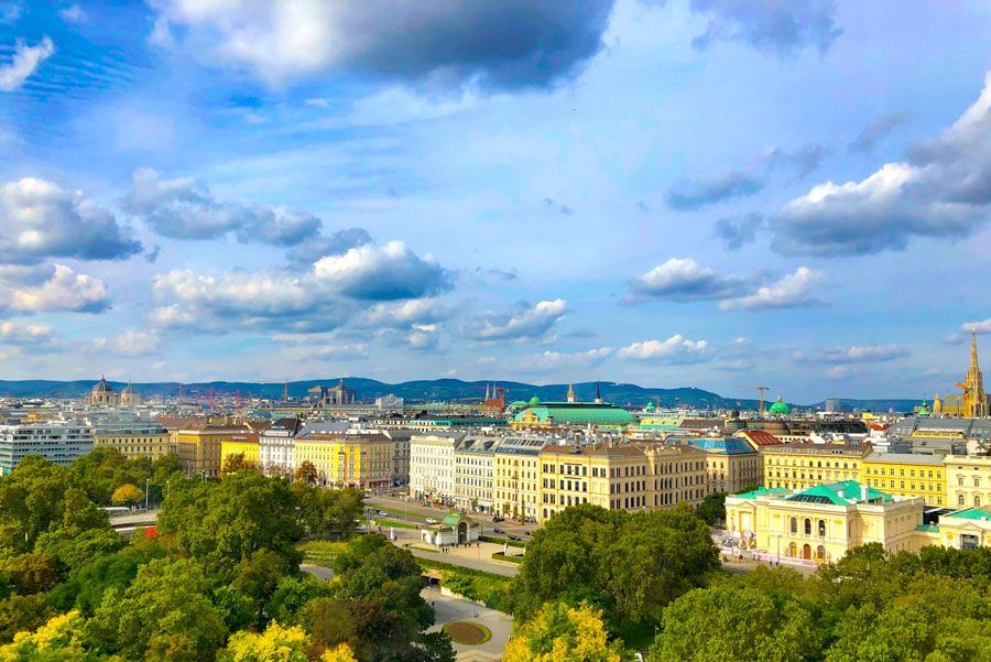 Paisaje urbano de Viena, Austria, bajo un cielo azul nublado. Edificios de diversos colores se alinean en el horizonte, con árboles y parques en primer plano.