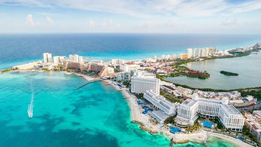 Vista aérea de Cancún, México, con océano turquesa, edificios blancos a lo largo de la costa y una laguna a la derecha.