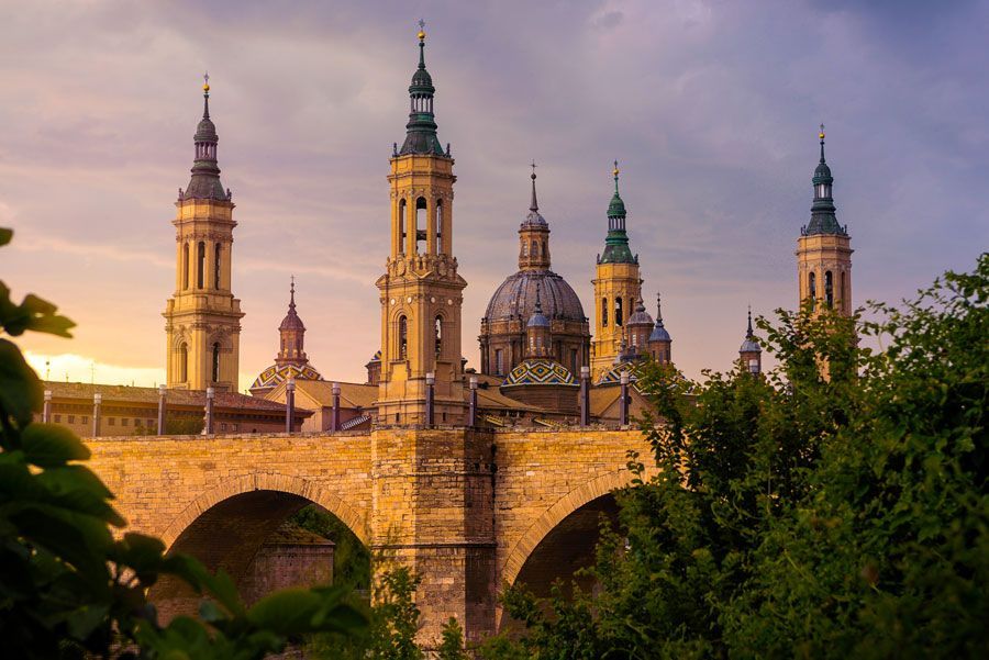 Basílica del Pilar en Zaragoza, España, al atardecer. La catedral, de piedra clara, cuenta con varias torres y cúpulas altas y ornamentadas que se recortan contra un cielo colorido.