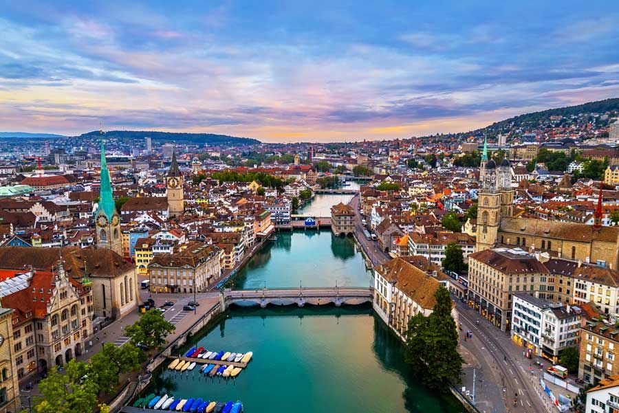 Vista aérea de Zúrich, Suiza, con un río que atraviesa la ciudad, rodeado de edificios con tejados rojos y barcos azules.