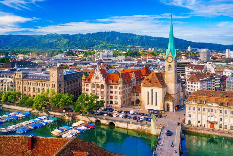 Vista aérea de Zúrich, Suiza, que muestra la ciudad con un río, barcos, edificios y una iglesia con una torre alta y verde.