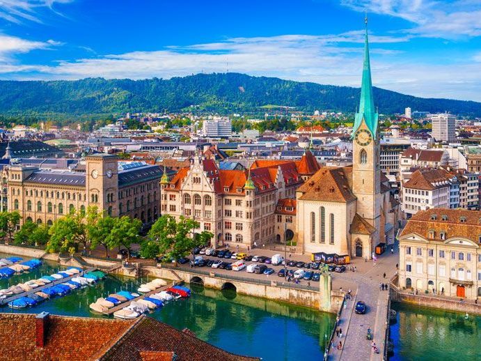 Vista aérea de Zúrich, Suiza, con un alto campanario de iglesia, edificios con techos rojos, un canal con barcos y un cielo azul con montañas al fondo.