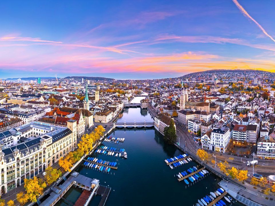 Vista aérea de Zúrich, Suiza, con un río que atraviesa la ciudad, barcos atracados, edificios y un cielo colorido al atardecer.
