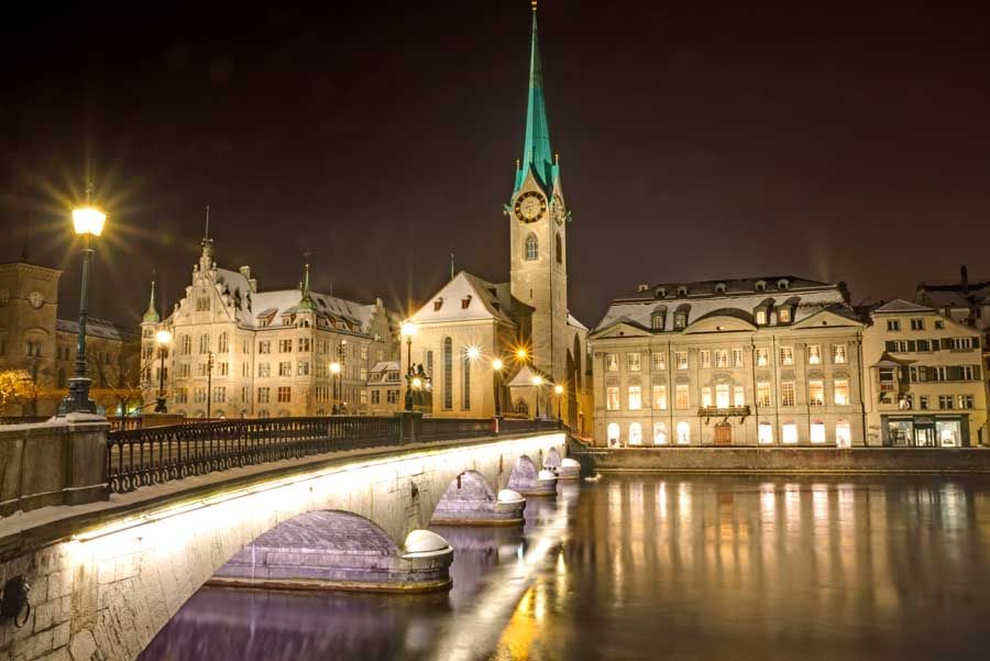 Vista nocturna de Zúrich, Suiza, con un puente cubierto de nieve, una iglesia con una aguja verde y edificios iluminados que se reflejan en el agua.