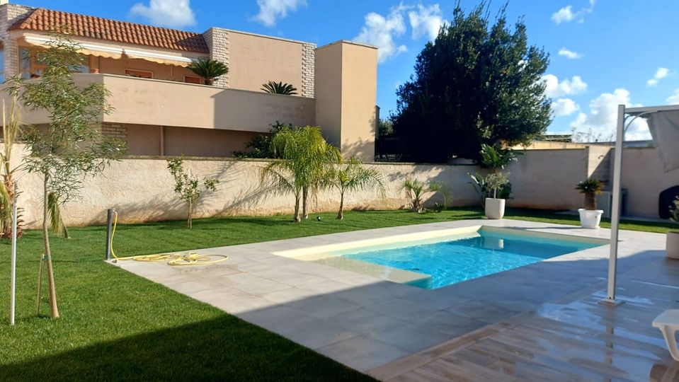 Moderno cortile con una piccola piscina blu, patio, prato e casa beige sotto un cielo soleggiato