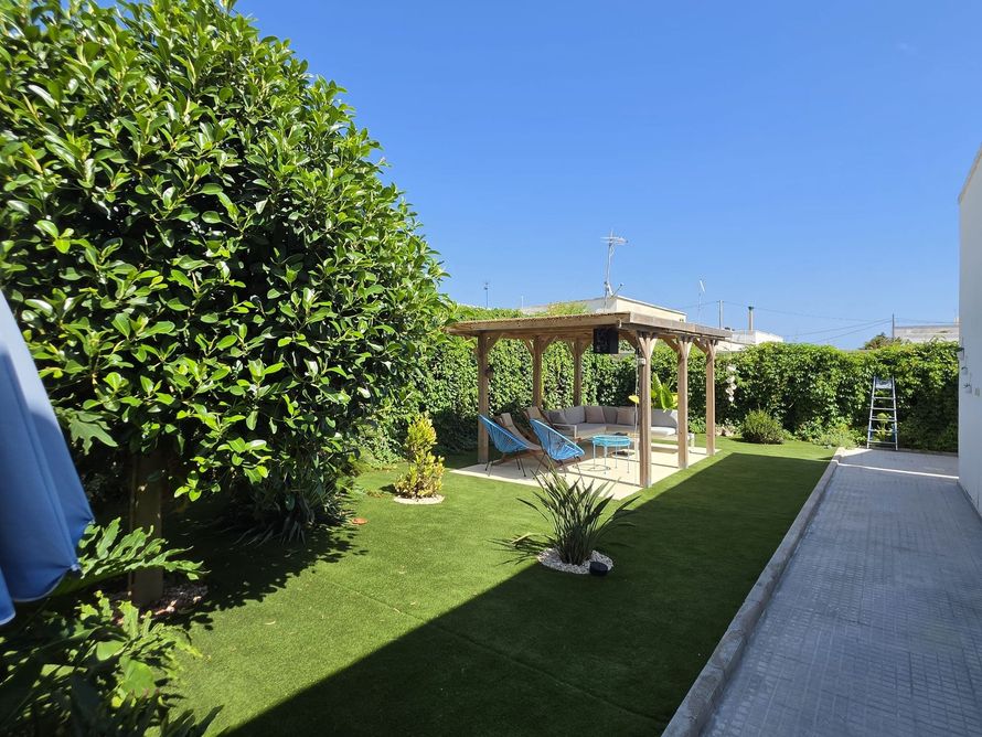 Giardino soleggiato con prato verde, pergolato in legno, siepi e un sentiero lastricato accanto alla piscina.