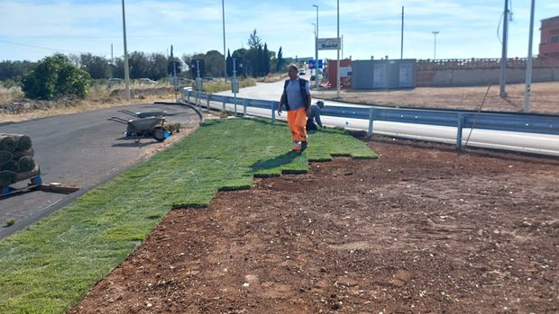 Un operaio posa del manto erboso lungo un'area di cantiere a bordo strada, accanto a tubi e terra.