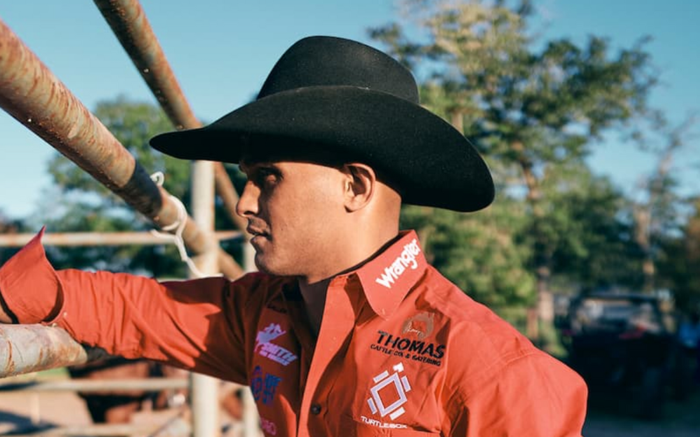 Man in cowboy hat leans on a fence, wearing a red shirt, looking away.