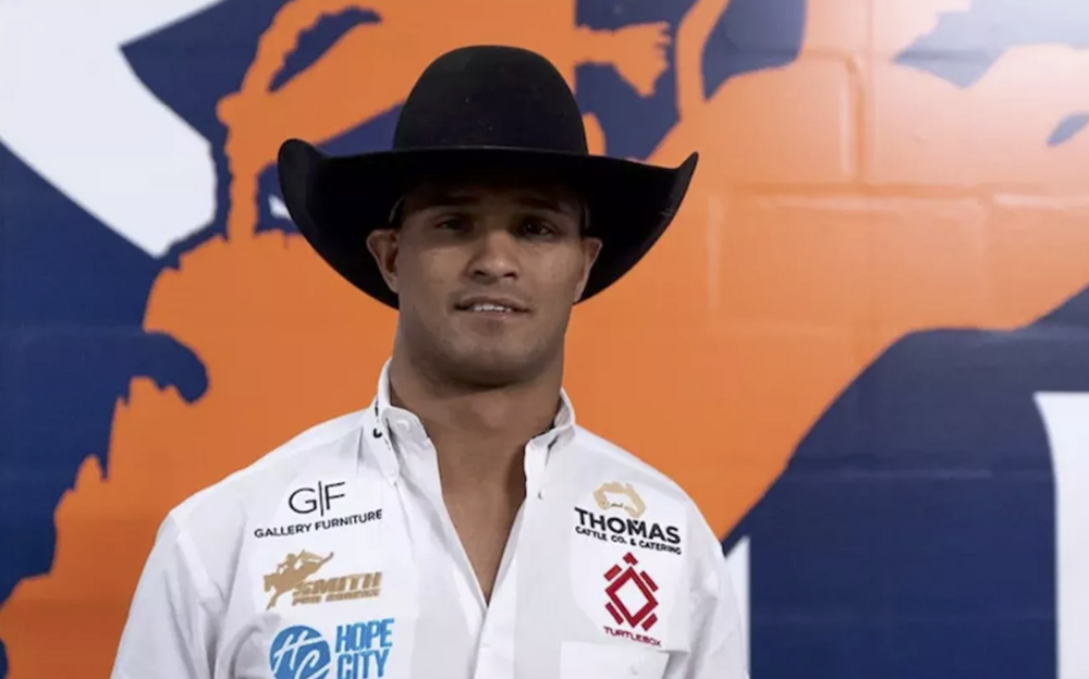 Man in cowboy hat smiles, in front of a blue and orange mural, wearing a white shirt with sponsor logos.