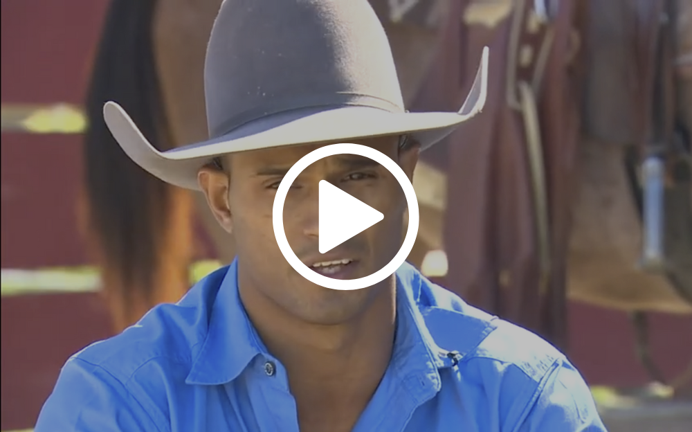 Cowboy in a blue shirt and grey hat speaking outdoors in front of a red fence and horses.