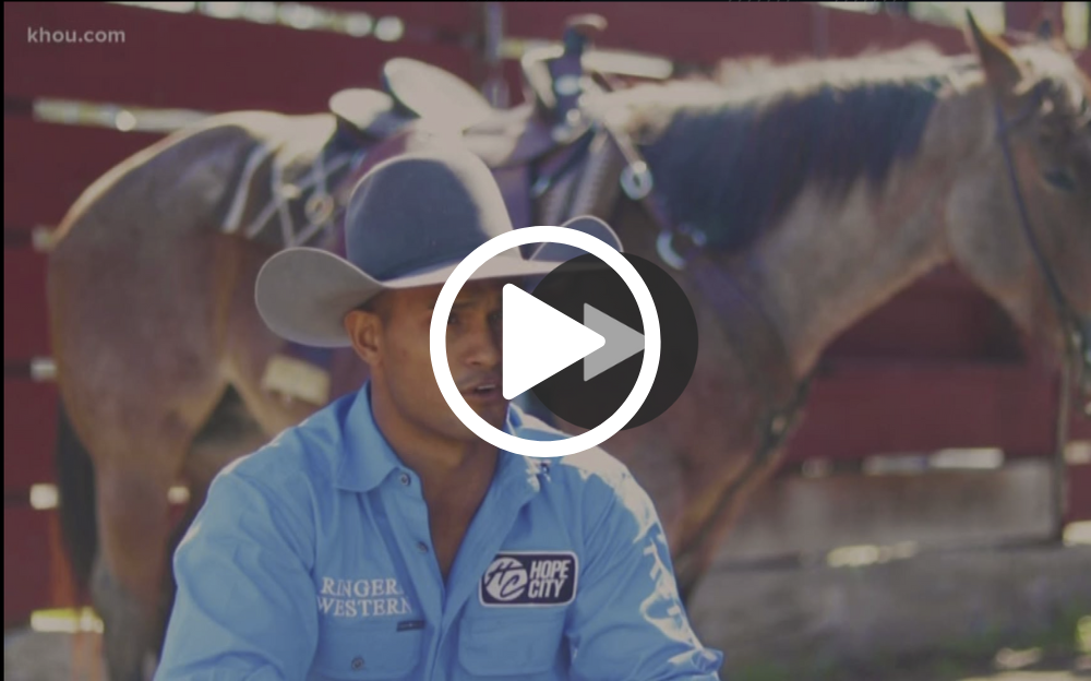 Cowboy in a blue shirt and hat, talking; horse with saddle in background. Outdoor setting.