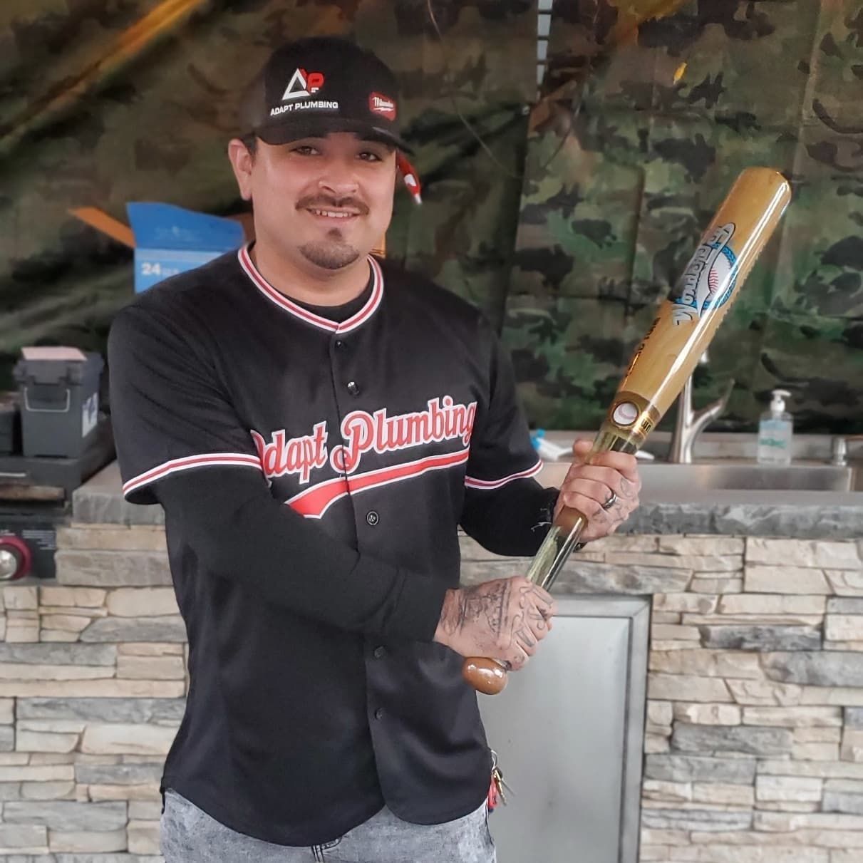 A man in a maple plumbing jersey holds a baseball bat