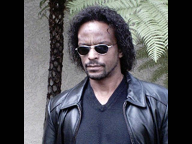 Man with sunglasses, dark hair, leather jacket, looking down, outside near plants.