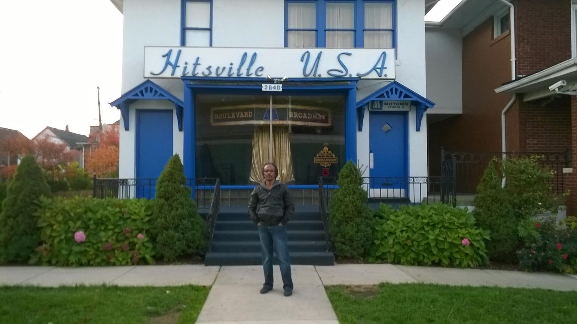 Man stands in front of Hitsville U.S.A. building; white and blue facade, with the words 