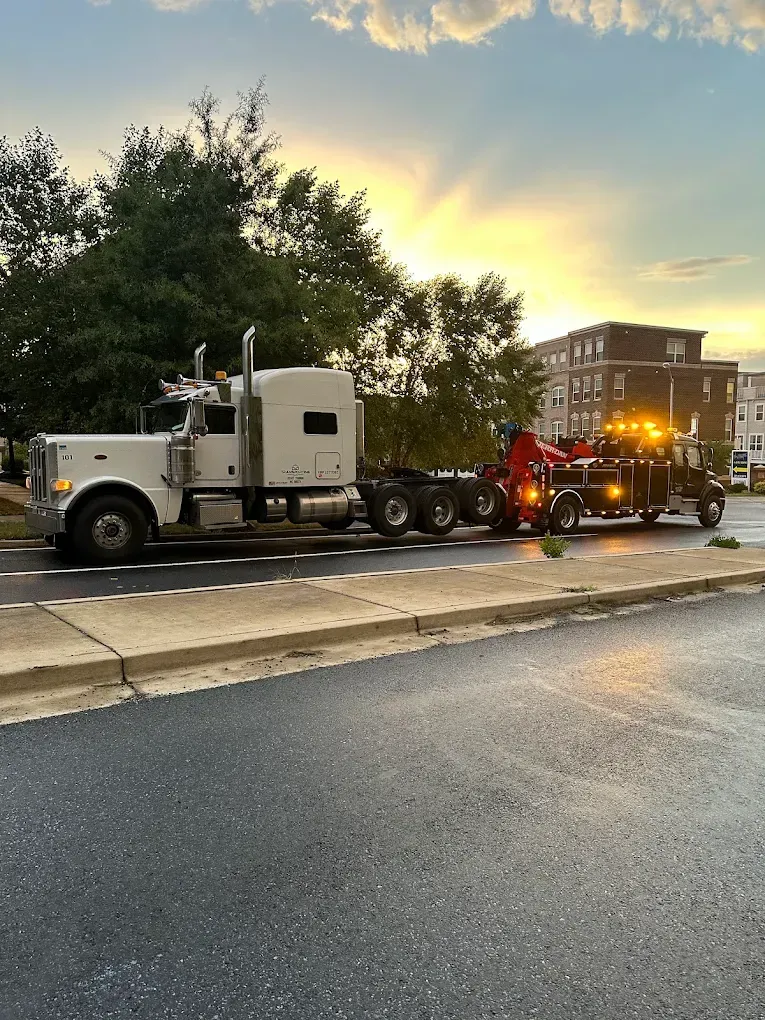 White semi-truck being towed by a tow truck on a wet road at sunset.