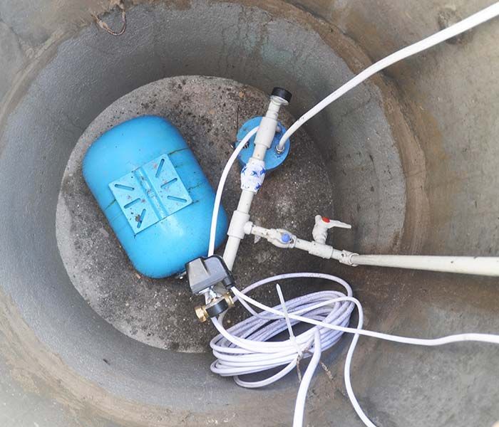 Blue well pump tank and attached white hoses inside a concrete underground pit.