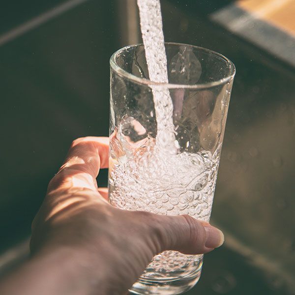 Hand pouring water into a clear glass held in a sunlit sink area