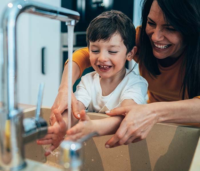 Parent and child washing hands together at a sink, smiling and playing with running water