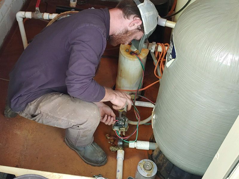 Technician crouched inside equipment bay, working on valves and hoses beside large tanks