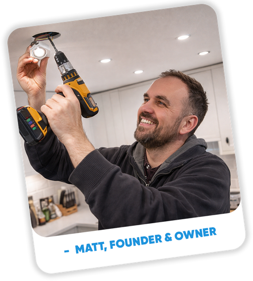 Man installing a light fixture with a power drill in a kitchen, smiling.