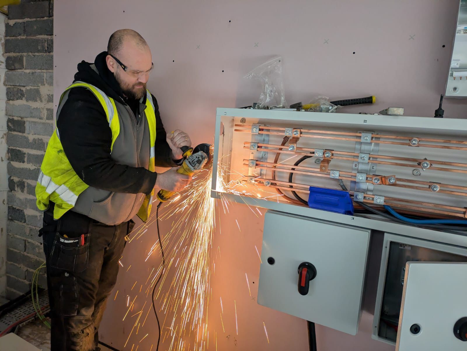 Construction worker using a grinder, creating sparks, on electrical panel in a building.