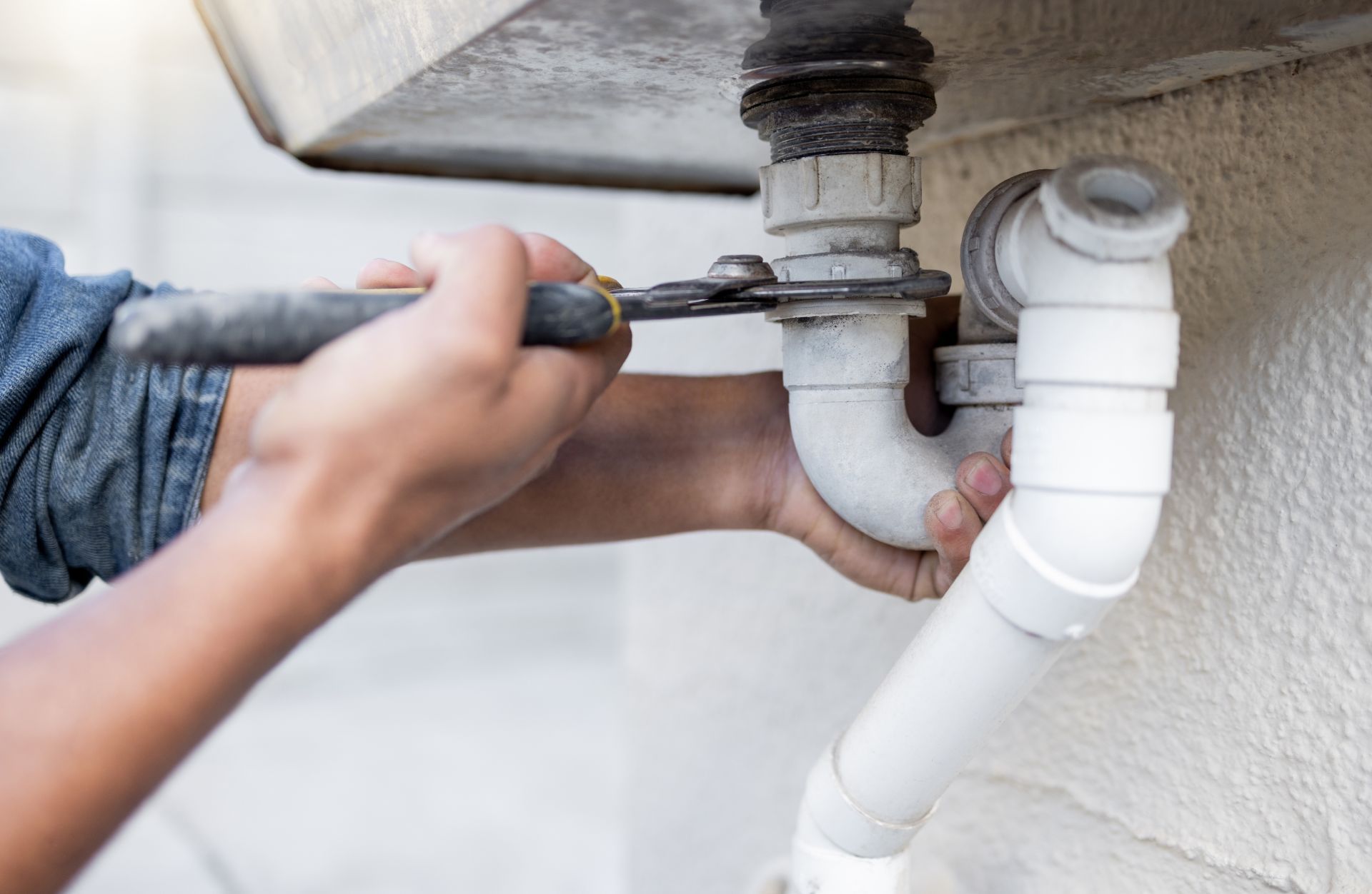 A person uses pliers to tighten a white plastic plumbing pipe under a sink.